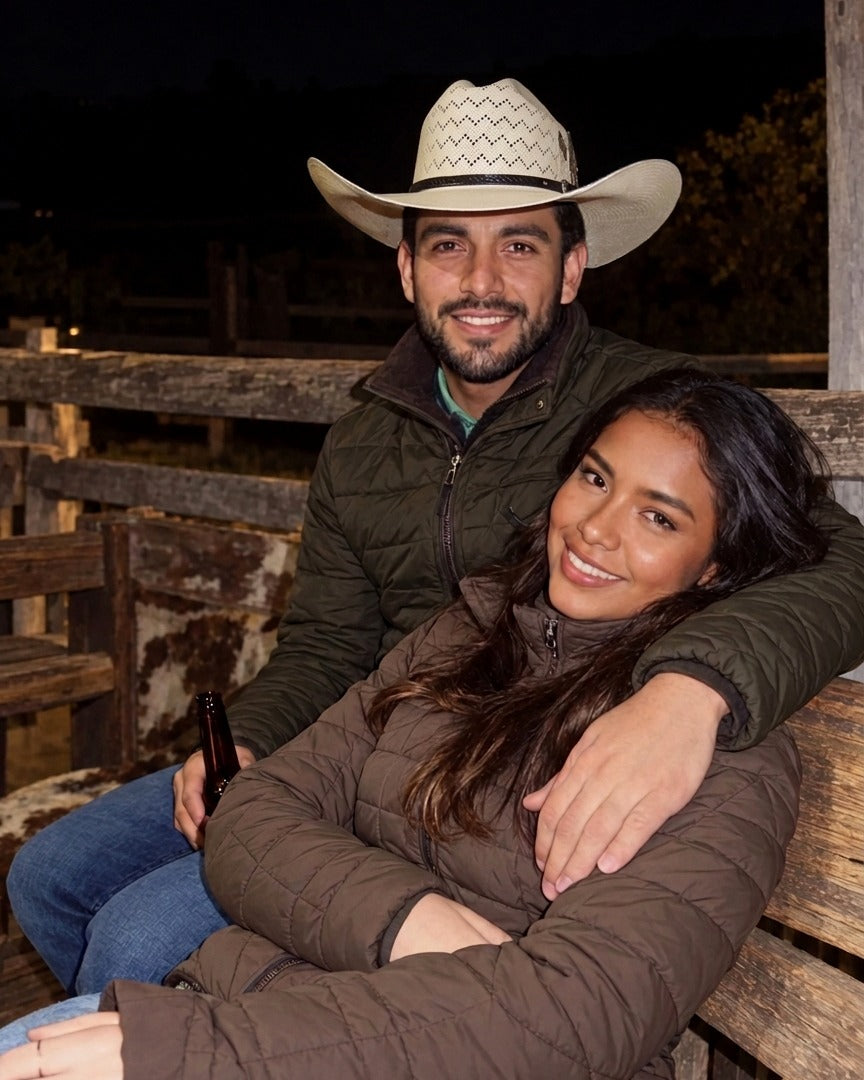 Pareja sonriente y abrazada en una noche de campo, sentada junto a una cerca de madera; él lleva un sombrero vaquero claro.